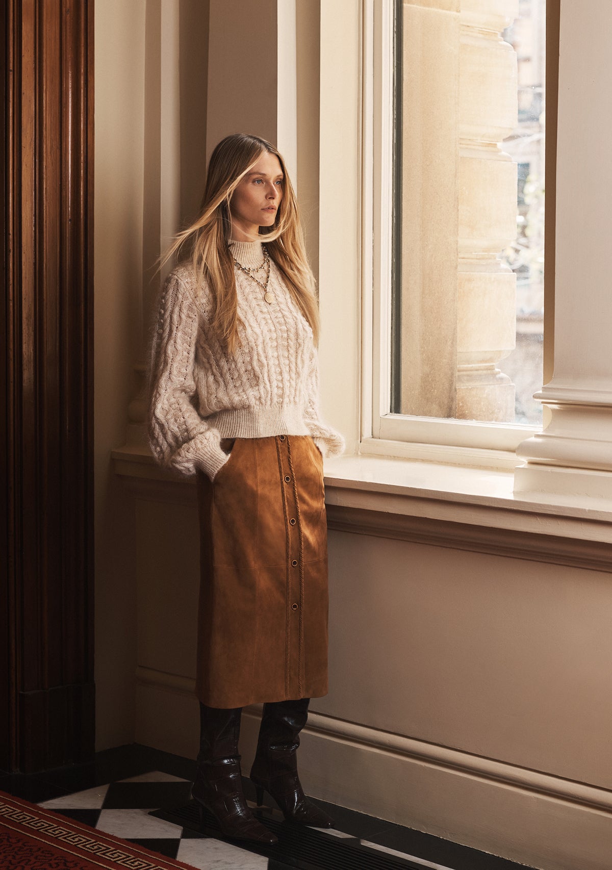 Woman in a patterned sweater and brown skirt standing by a window in a room with checkered floor.