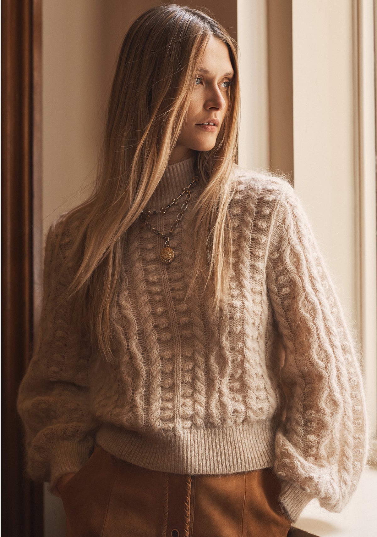 Woman wearing a beige knitted sweater standing by a window.